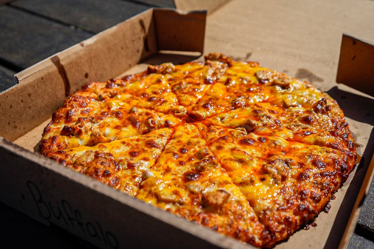 A freshly baked rectangular pizza in a cardboard box, topped with melted cheese and scattered toppings, sitting in bright sunlight