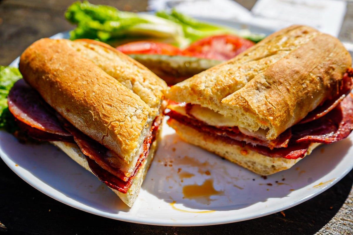 Two crusty sandwiches with sliced cured meat on a white plate, surrounded by fresh vegetables including lettuce and tomato