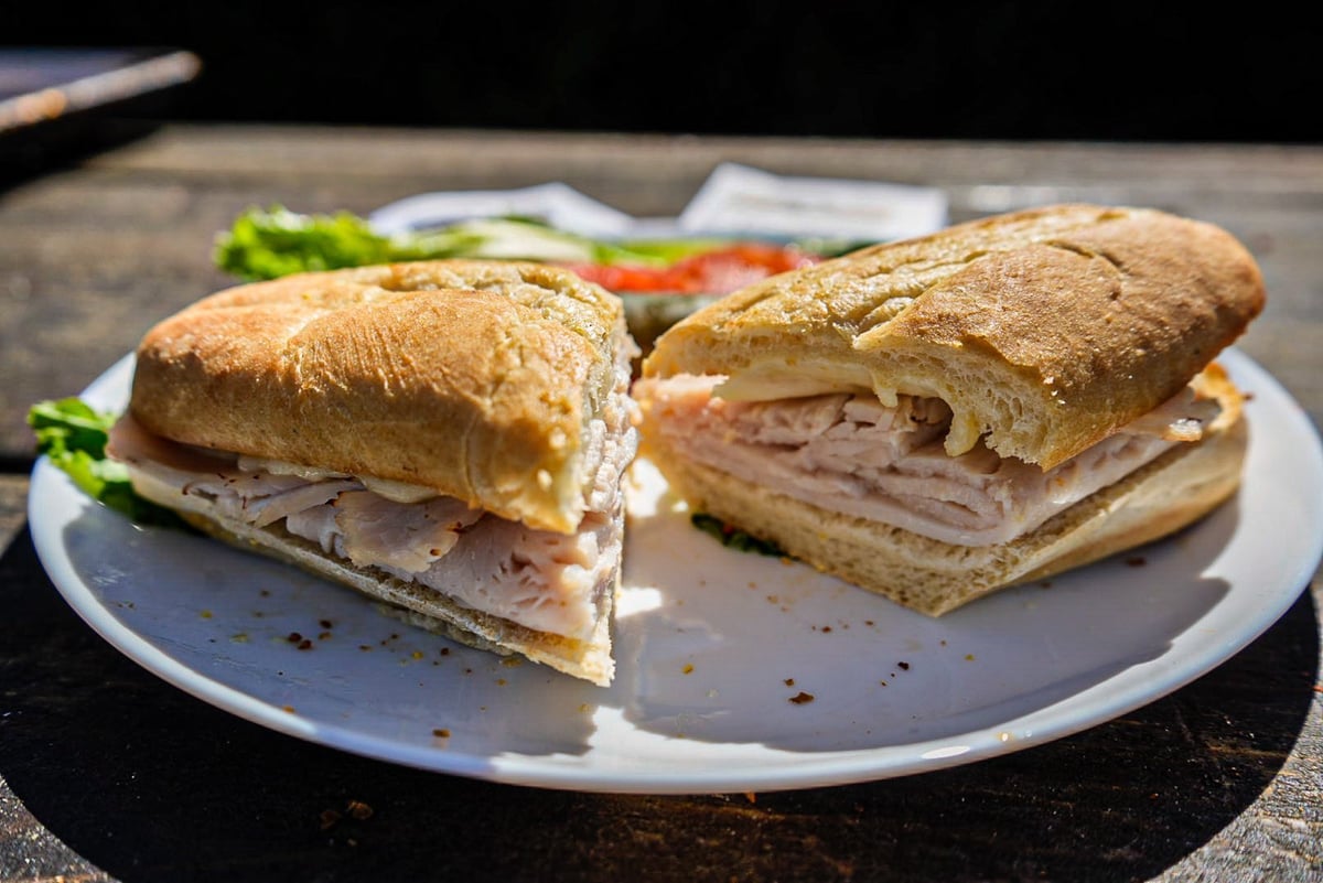 Two halves of a sliced sandwich with meat filling served on a white plate with lettuce and tomato in the background