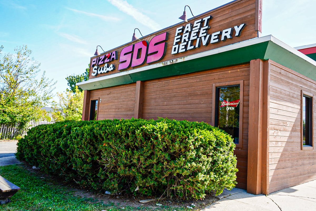 Retro SDS Pizza and Subs restaurant exterior with pink neon sign, brown wooden siding, and green shrubs in front