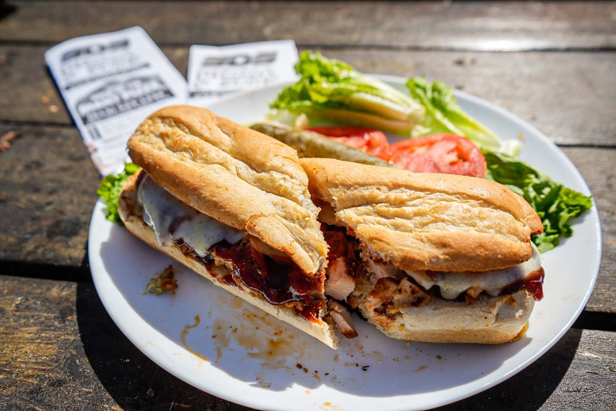 Crusty sandwich with meat and sauce served on white plate with lettuce and tomato garnish on wooden table