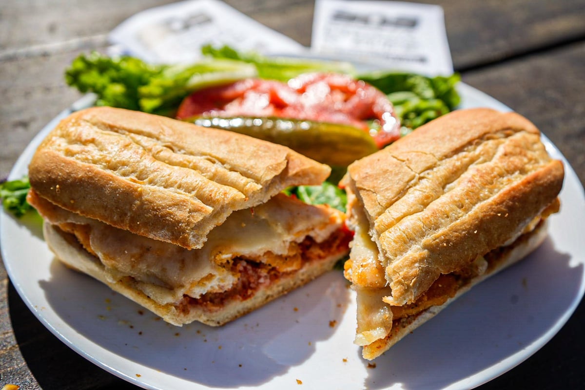 Cuban sandwich cut in half on a white plate, showing ham, cheese, and pickles, with salad and side items in background