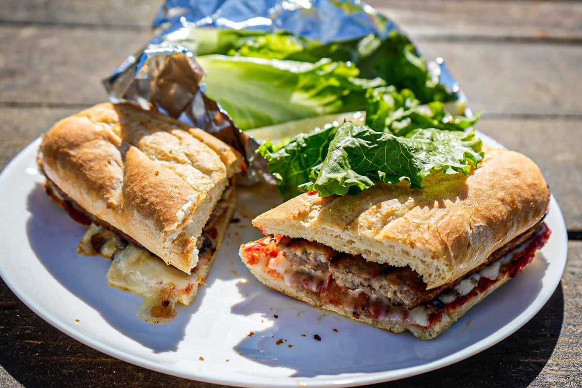 Two crusty sandwiches with tomato and meat filling served on a white plate with fresh green salad on a wooden table