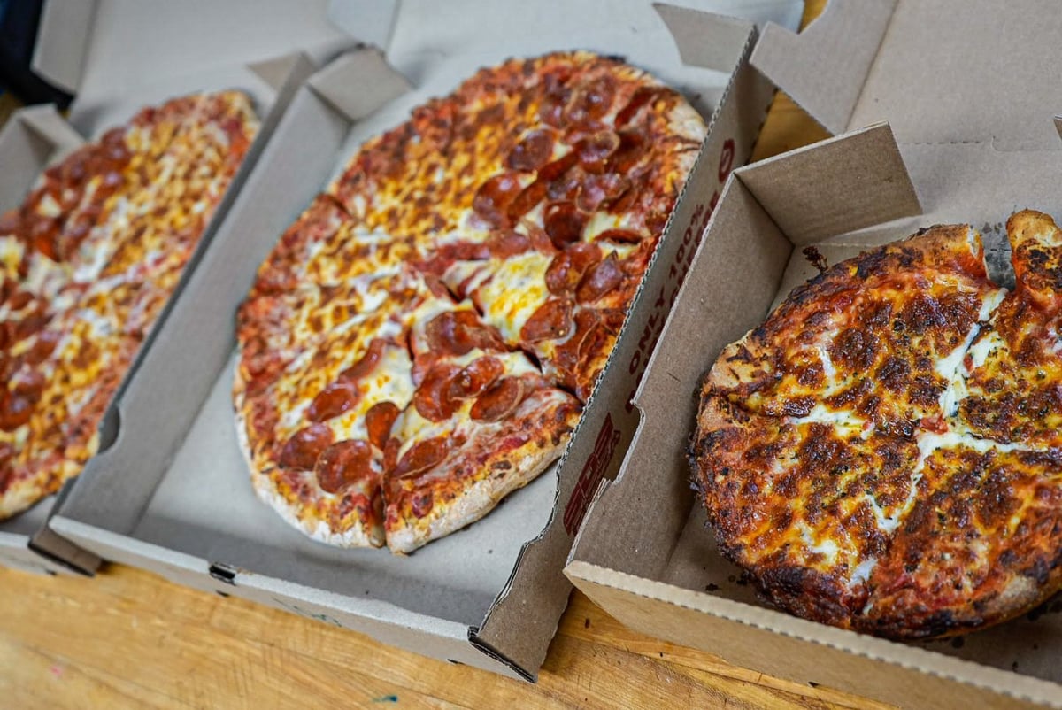 Three pizzas in cardboard boxes arranged on a wooden surface, featuring pepperoni and cheese toppings with browned melted cheese