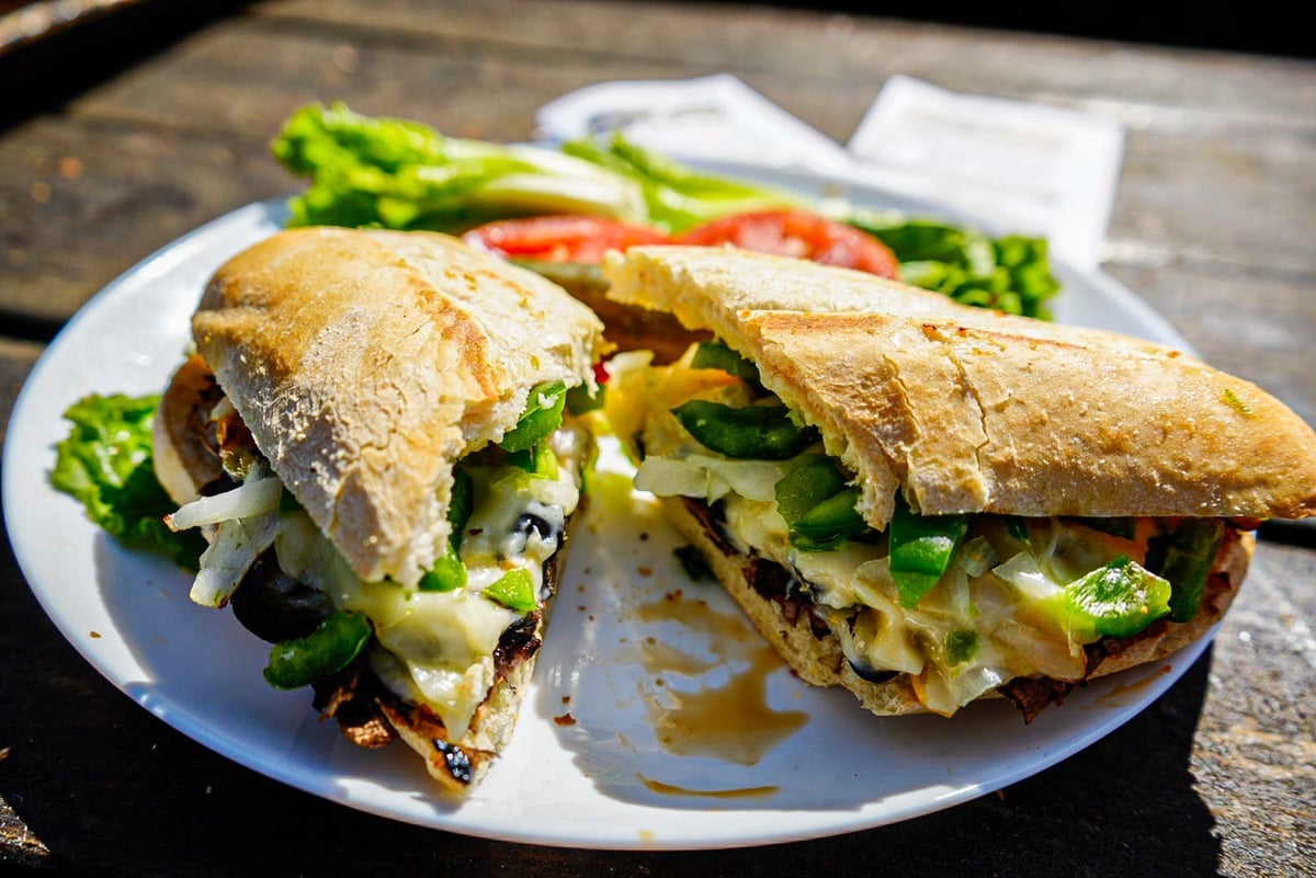 Two crusty baguette sandwiches with melted cheese, lettuce, and other fillings on a white plate with fresh vegetables in the background